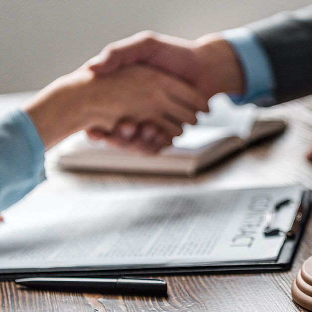 Zwei Personen sitzen sich am Tisch gegenüber und schütteln sich die Hände, auf dem Tisch liegen Papiere und ein Richterhammer - Gesetze beim Hausverkauf